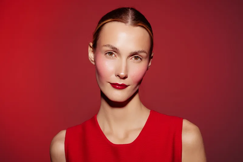 Portrait of a woman with red lipstick and blush wearing a red sleeveless top against a red background. Concept of fashion, beauty, makeup, and elegance in studio lighting.