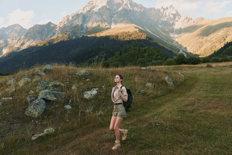 Young woman hiking across a mountain meadow with a backpack, gazing toward distant peaks and enjoying nature, sunlight casting long shadows over rugged slopes.