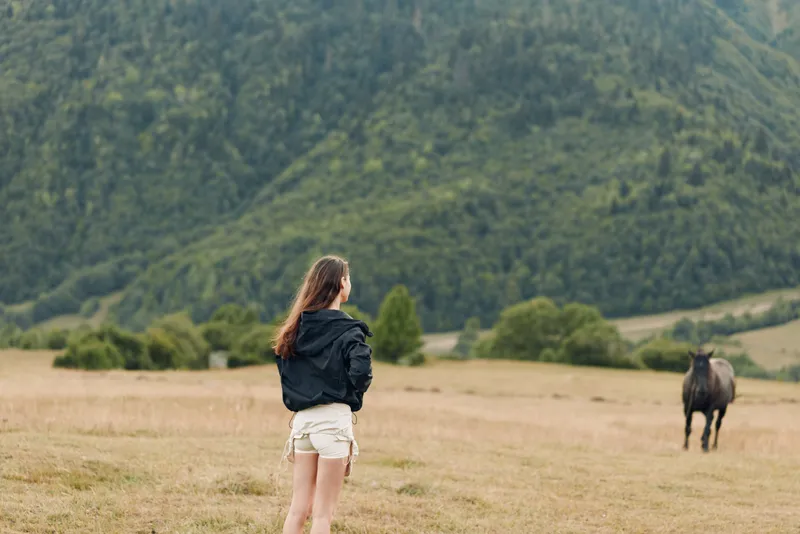 A young woman stands in a wide open field, watching a horse in the distance. She wears a black jacket and light shorts, enjoying the tranquil countryside and fresh air.