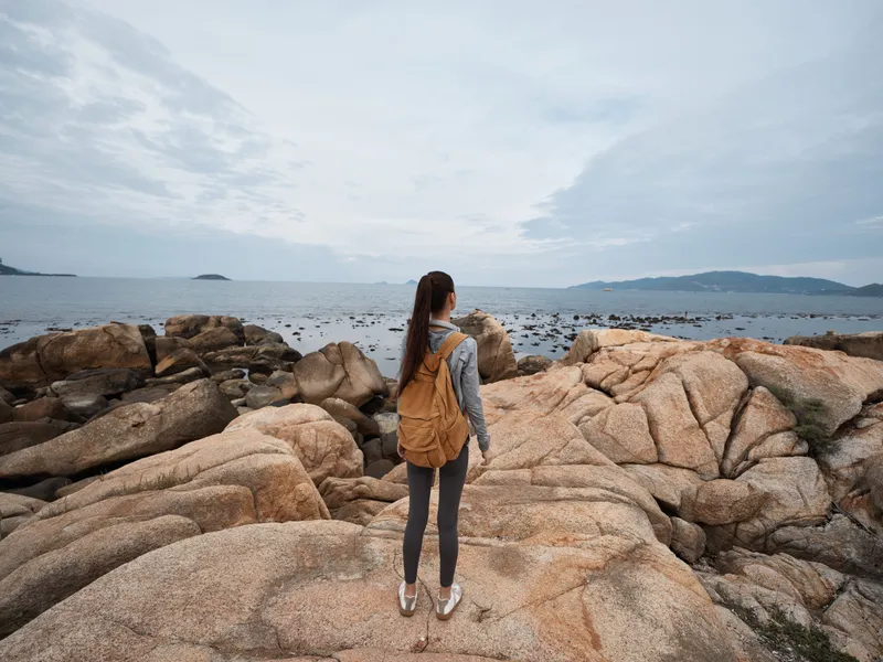 Woman with a backpack enjoying serene ocean view on rocky beach, wearing casual outfit, reflecting calm emotions in natural scenery