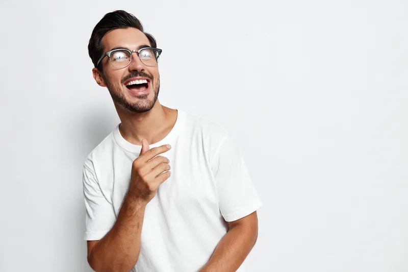 Smiling young man wearing glasses and a casual t-shirt, looking at the camera with a joyful expression while posing against a solid light background.