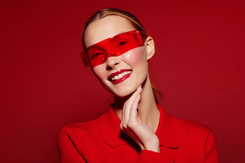 Smiling woman with red sunglasses and red outfit poses against red background. Fashion portrait with bright makeup and confident expression. Concept of style and modern beauty.