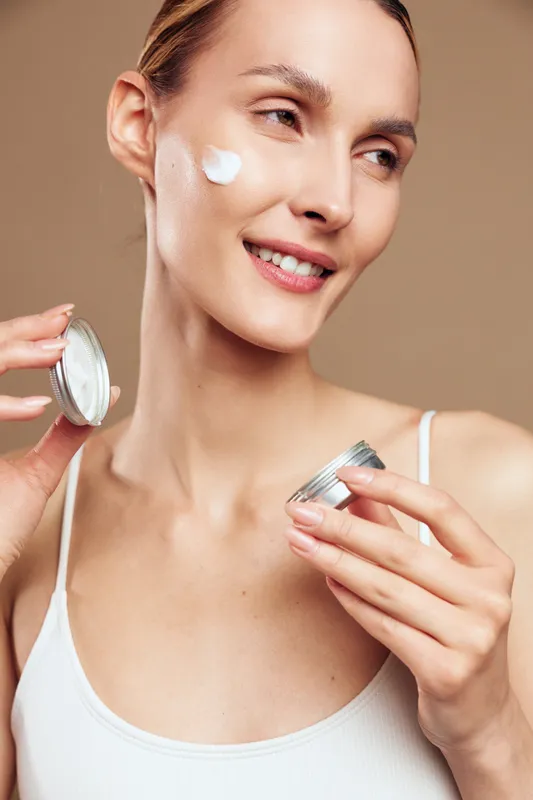Young woman applying face cream, smiling gently, natural skin care routine, soft lighting, beige background, casual white top, close up portrait, healthy glowing complexion