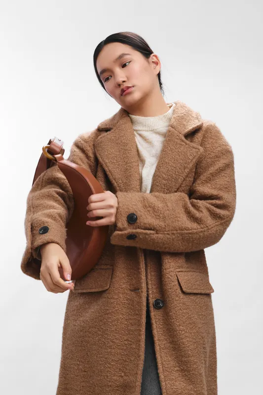 Asian woman wearing a warm, oversized brown coat adjusts a round leather bag, posing confidently in a bright studio. The clean backdrop emphasizes her style and poised presence.