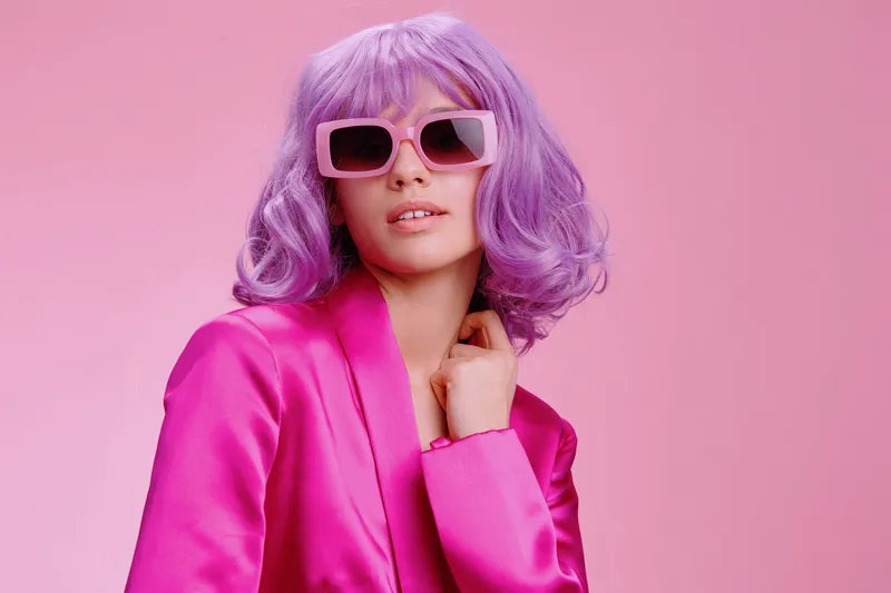 Женщина с фиолетовыми волосами в солнцезащитных очках и розовом костюме стоит на розовом фоне, демонстрируя смелое и стильное модное заявление благодаря яркому цвету волос и подходящему наряду