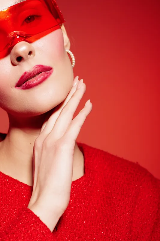 Close-up of stylish woman in red sweater and red sunglasses posing with hand near face on red background. Fashion portrait with bold makeup and modern accessories.