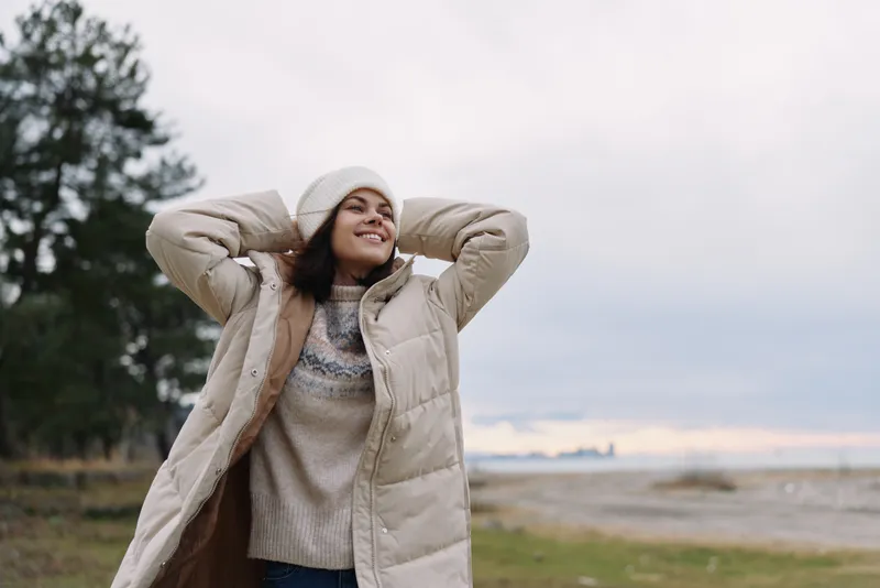 Woman outdoors in a warm beige coat and knit sweater, smiling with hands behind head, autumn breeze by a sandy shoreline and trees at a quiet coast, peaceful mood and carefree style
