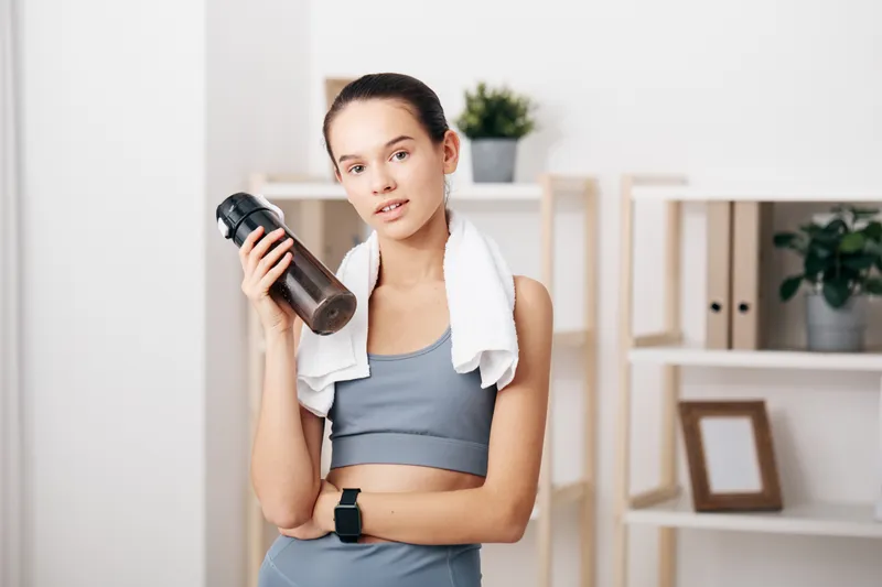 woman fitness sport workout training health wellness athletic exercise indoors, a female athlete stands with a water bottle and towel in a bright living space, ready for a home workout and daily