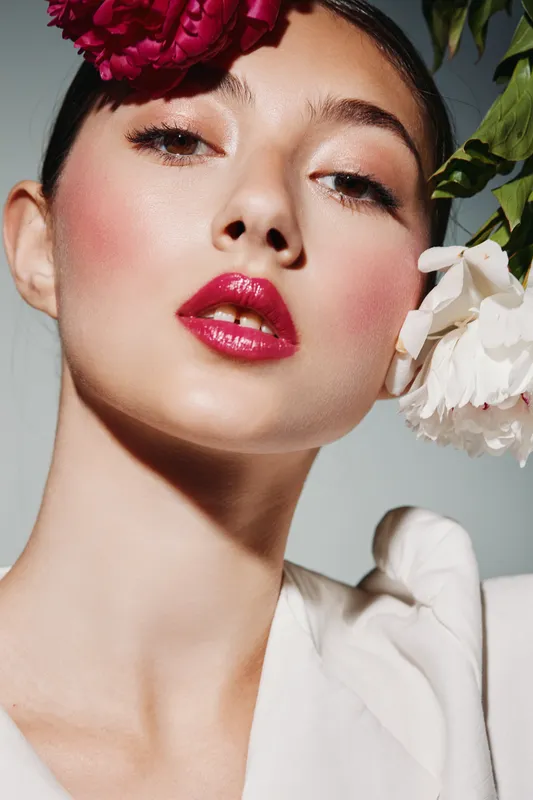 Close-up of woman with creative makeup, glossy red lips, and blush, wearing white garment with flowers threading her hair. Concept of beauty, fashion, and floral elegance in studio.