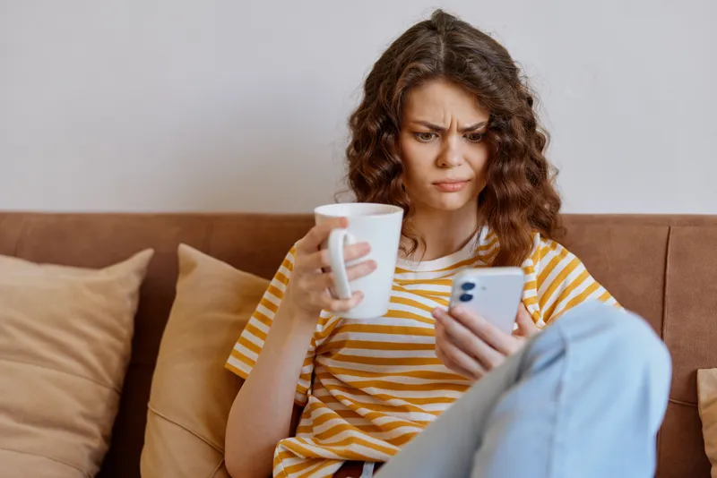 Casual woman at home, sitting on a sofa, sipping from a mug while looking at a smartphone, relaxed lifestyle moment, striped tee, soft natural light, cozy domestic setting