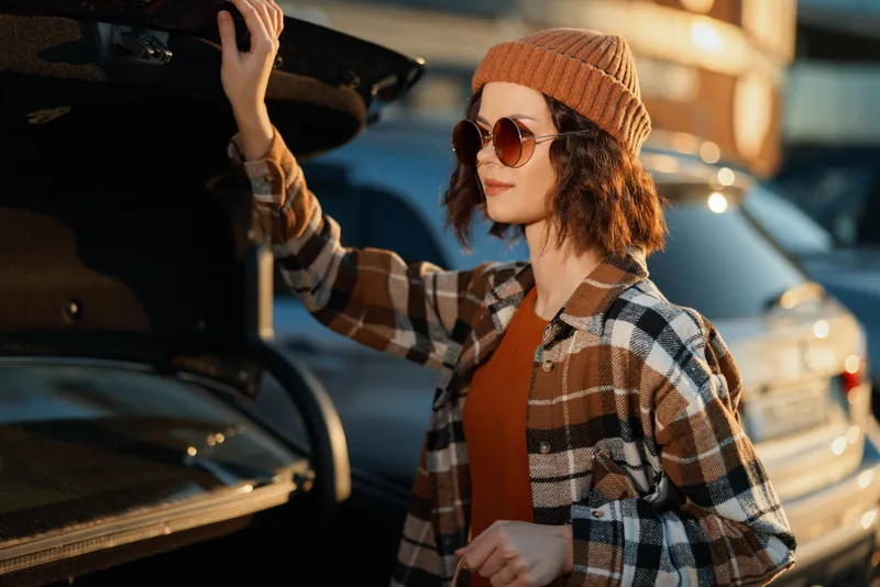 woman car trunk sunglasses beanie plaid golden sunlight Young woman opens a car trunk at golden hour wearing sunglasses and beanie in a plaid shirt; candid lifestyle scene with authenticity and