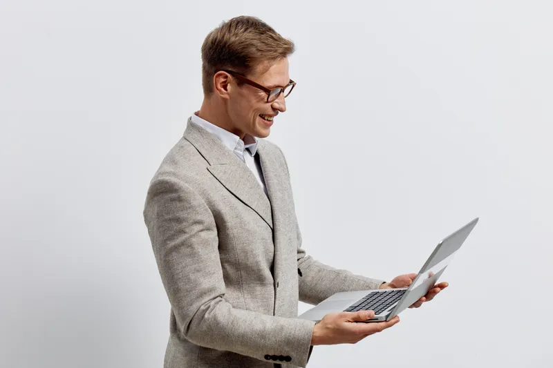 Smiling man wearing glasses, beige blazer, white shirt, using a laptop, standing against plain light background. Business casual, technology, work, professional, office, communication style.