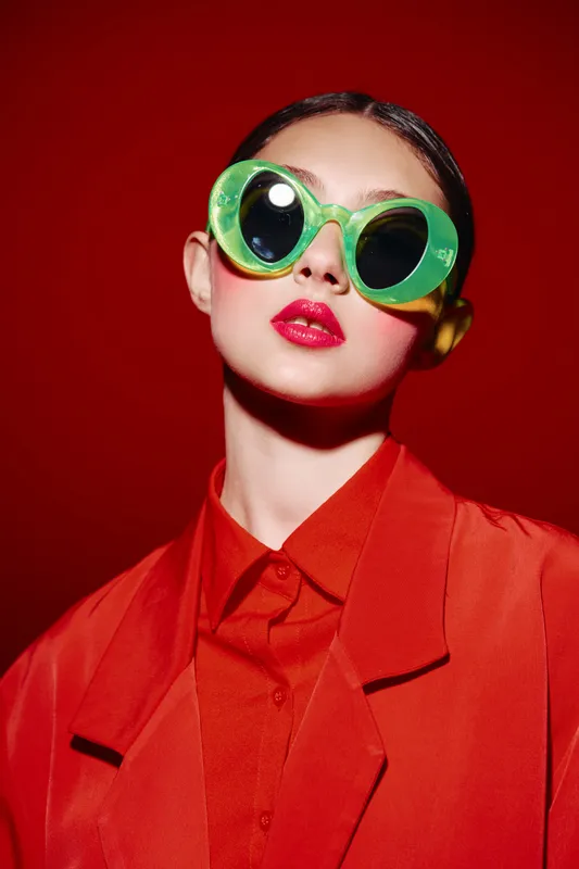 Stylish young woman wearing bright green oversized sunglasses and intense red lipstick posed against a vibrant red background. Concept of bold beauty, fashion creativity.