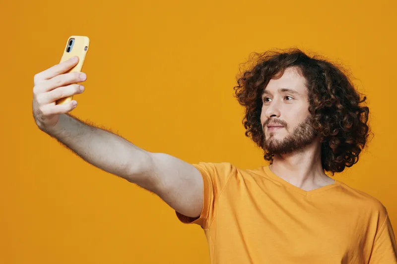 Young man with curly hair taking a selfie against a vibrant yellow background, capturing a joyful moment while dressed in a casual orange t shirt
