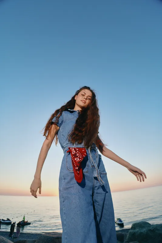 lifestyle woman in modern boho-western denim street style with long hair and red bandana by the sea at sunset, film color tones evoke relaxed outdoor mood