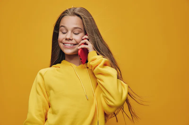 Young girl with long hair wearing yellow sweater smiling with eyes closed while talking on red phone against yellow background. Happy communication, lifestyle and technology concept.