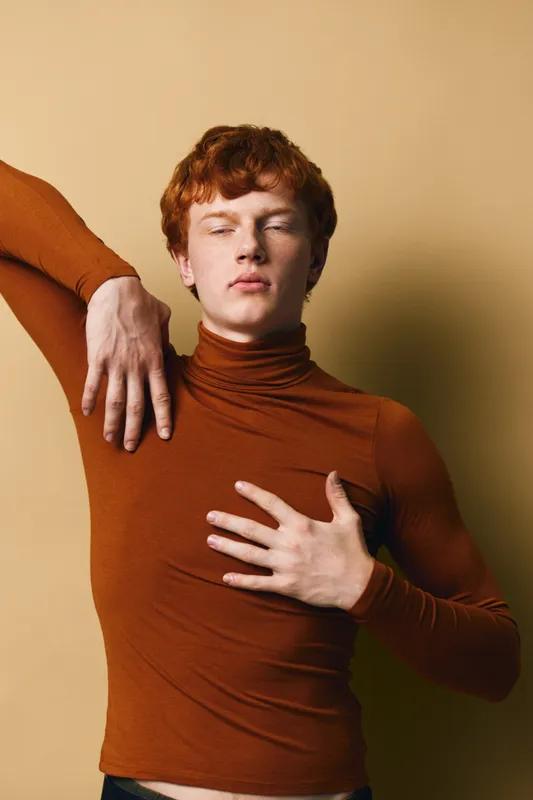 Young man with red hair wearing a brown turtleneck poses with eyes closed and hand on chest against beige background. Fashion, portrait, expression and style in studio lighting.