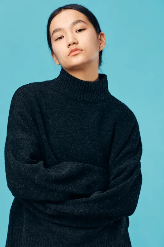 Young woman with long dark hair wearing black knit sweater standing with crossed arms against blue background, looking confident and serious in casual fashion portrait.