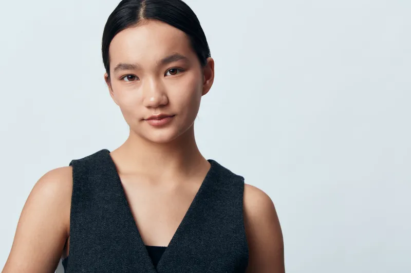 Young woman with dark hair and clear skin wearing sleeveless black top posing with neutral expression on light background. Portrait, beauty, fashion, confident, studio lighting.