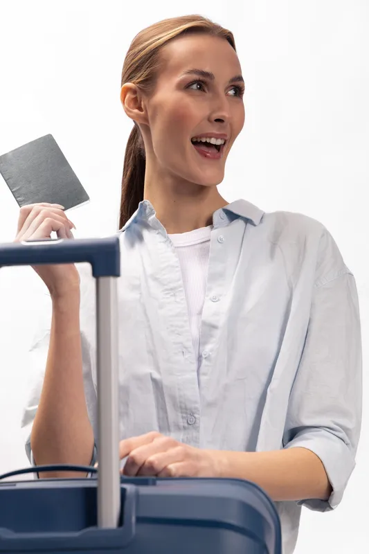 Happy woman in casual shirt holds passport and stands behind blue suitcase. Concept of travel, vacation, tourism, and holiday planning with excitement and positive mood.