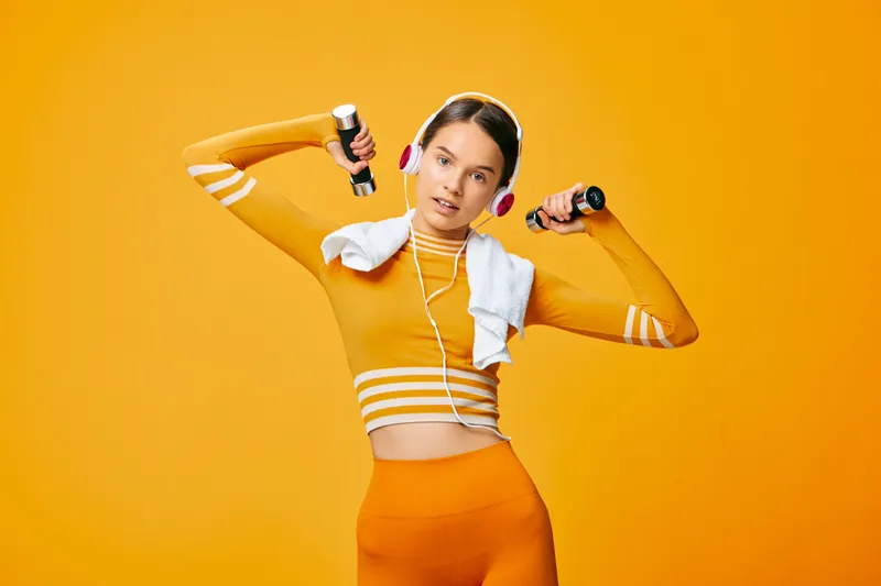 Fit woman in orange activewear lifts small dumbbells while wearing headphones and a towel, studio fitness portrait on vibrant orange background conveying strength and confident energy.