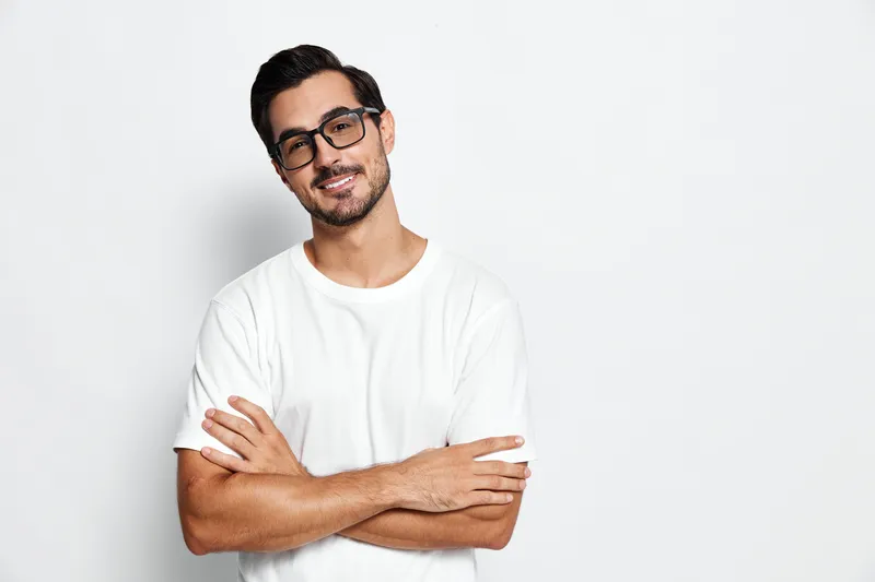 Smiling man with glasses standing confidently with crossed arms, wearing a casual white t-shirt against a plain light background. Portrait lifestyle concept.