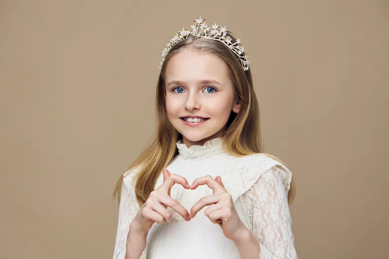 Portrait of a cheerful young girl in a white dress and tiara, making a heart shape with her hands against a warm beige background, capturing joy and innocence