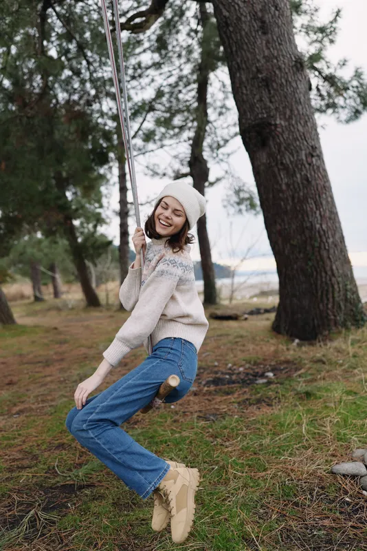 Girl on a swing in a park, smiling and enjoying outdoor leisure in a cozy sweater and boots. A candid moment among trees and grass, capturing natural lifestyle and playful outdoor activity.