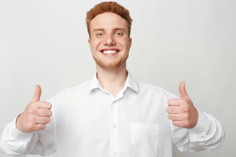 Smiling young man with red hair wearing white shirt shows thumbs up gesture with both hands. Positive expression on light background. Concept of approval, success, and confidence.