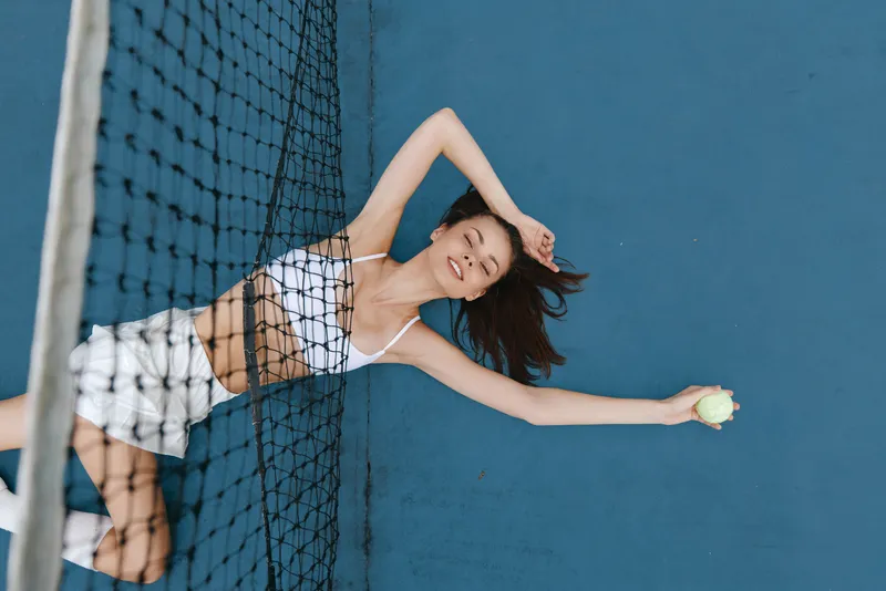 Young female tennis player in sporty attire smiling with a tennis ball on a blue court, capturing joy and confidence in an active lifestyle