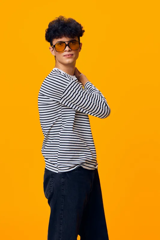 Young man wearing striped shirt and sunglasses poses confidently against vibrant yellow background, showcasing casual style and modern fashion.