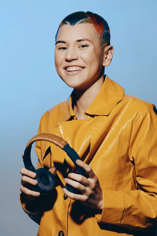 Smiling woman with multicolored hair wearing a bright yellow jacket holds black headphones against a soft blue background with confident and joyful expression.