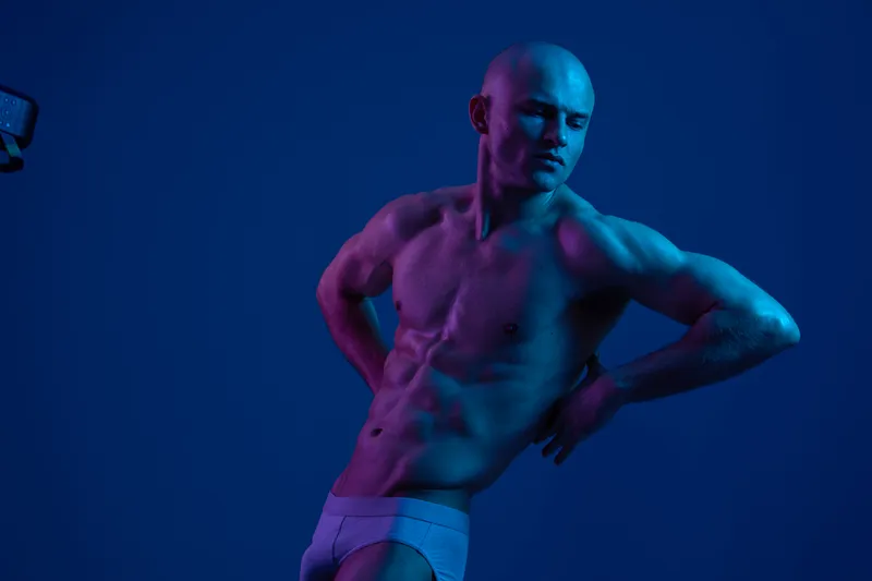 Athletic man posing confidently in a modern blue-themed studio, showcasing strength and elegance. The striking lighting enhances the body's contours and forms.