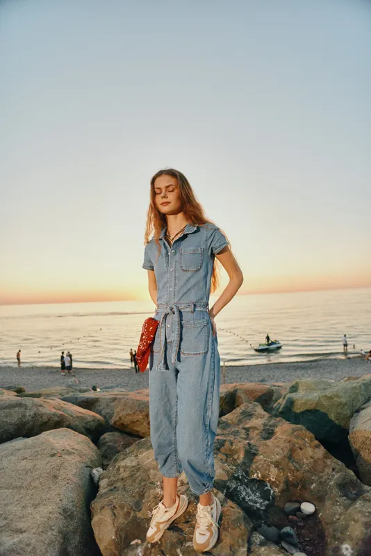 Lifestyle woman in modern boho-western denim street style stands on rocks by the sea at sunset with relaxed pose and film color tones enhancing mood.