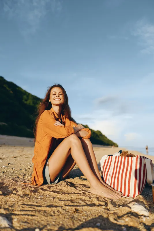 Bright, relaxed portrait of a smiling woman on a sunny beach, sitting on sand with striped tote, orange shirt, enjoying outdoor summer mood and peaceful seaside scenery with warm light.