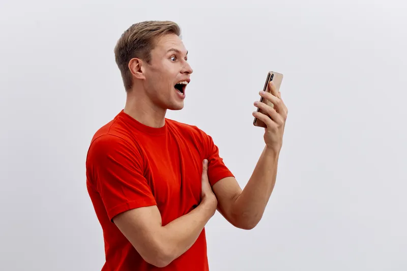 Smiling young man wearing red t-shirt looking at mobile phone with excitement isolated on a plain white background studio portrait. People lifestyle concept