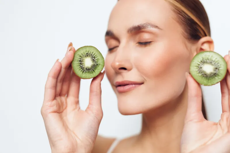 Young woman with smooth skin holds two halves of a kiwi fruit near her face with closed eyes and a gentle smile on a white background.