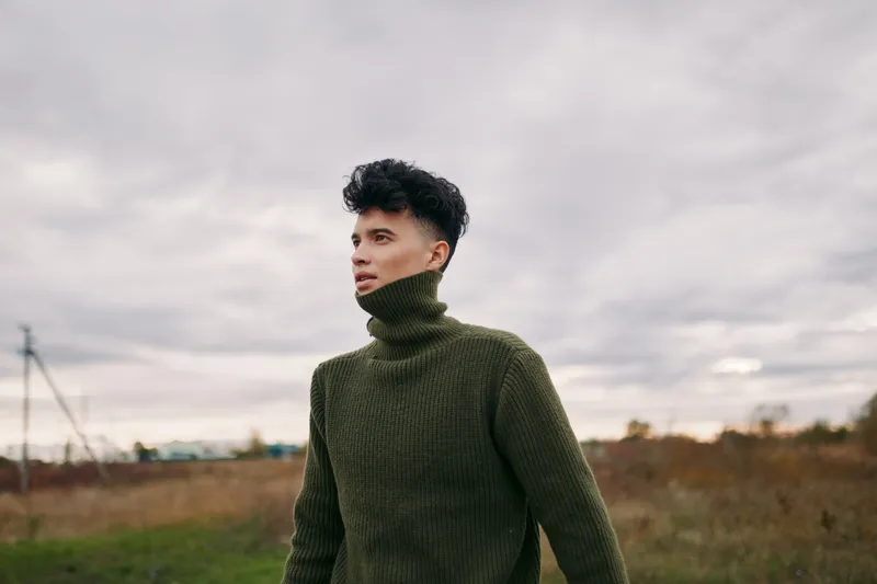 An authentic portrait of a young man in a chunky green turtleneck standing in an open field under a moody sky, conveying sincere presence and veracity.