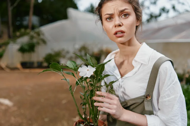 Young woman expressing concern while holding a potted plant in a greenhouse setting, showcasing emotional engagement with nature and gardening