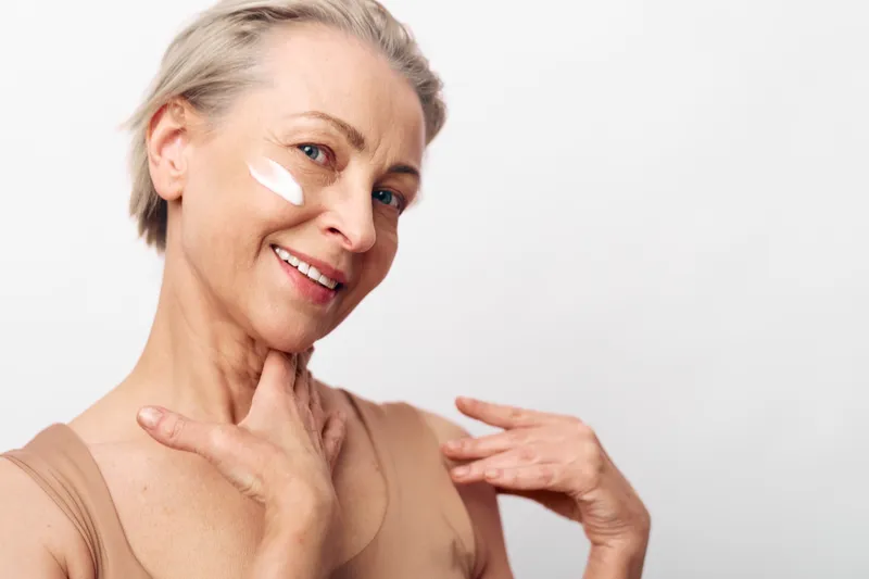 Smiling mature woman applies white face cream on cheek, showcasing skincare routine and healthy skin. Concept of beauty, self-care, aging gracefully, and wellness in natural light.