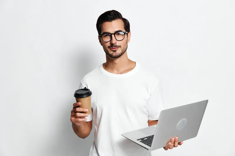 Confident smiling man in casual white t-shirt holding laptop and coffee cup while looking at the camera against a plain light background. Modern lifestyle concept.