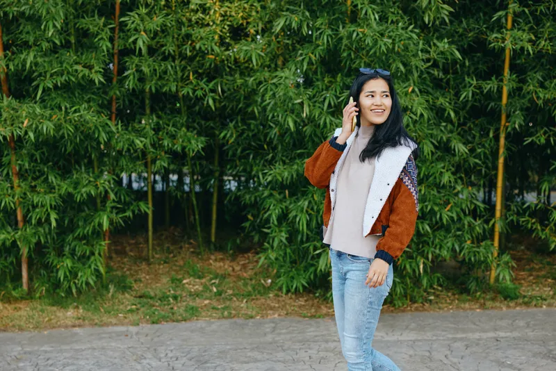 Woman on outdoor street talks on phone, wearing jacket and jeans, smiling amid greenery and bamboo, casual fashion vibe in natural daylight and urban ambiance