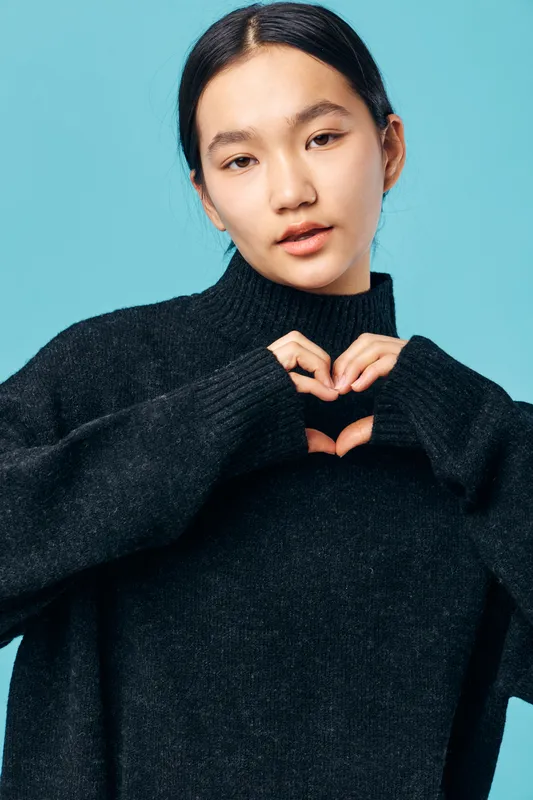 Young woman in black sweater making heart shape with hands against blue background, casual style, portrait with natural expression and modern fashion concept for love or care.