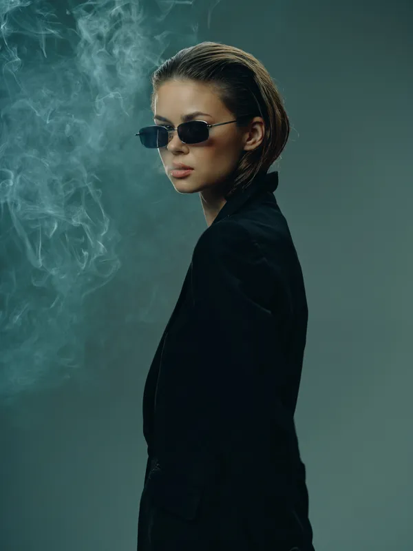 Stylish woman in sunglasses wearing black suit jacket with smoky background, confident pose and moody lighting.