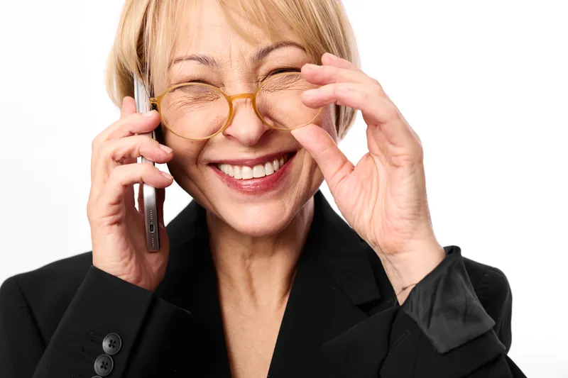 Senior caucasian woman in business suit smiles on phone call, adjusting glasses with confident friendly expression, studio portrait isolated on white background