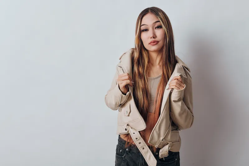 Young woman with long hair wearing beige jacket and casual clothes standing against white background looking confident with stylish expression and posture.