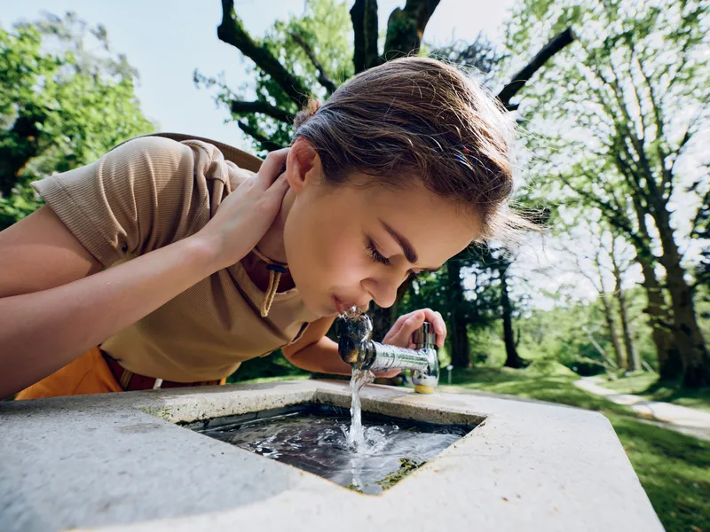 woman drinking fountain park water outdoors, young female leaning over public tap, refreshing hydration moment in green park with trees, sunlight and natural summer atmosphere