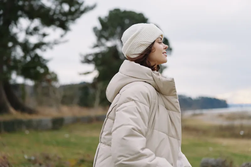 Woman in a beige coat and knit beanie stands outdoors by a lake with trees and cloudy sky, showcasing peaceful winter style and natural scenery, calm mood and seasonal fashion