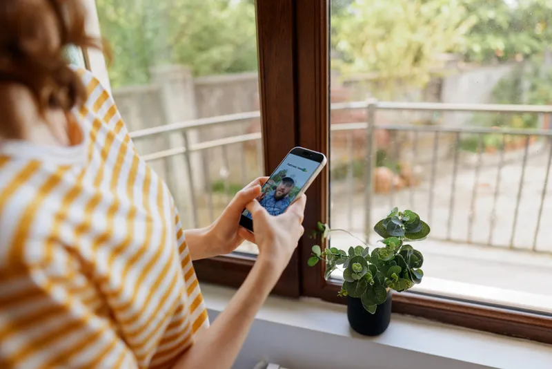 Young woman sits by a window scrolling on her phone, striped top, a cozy home moment with natural light and a small plant on the sill