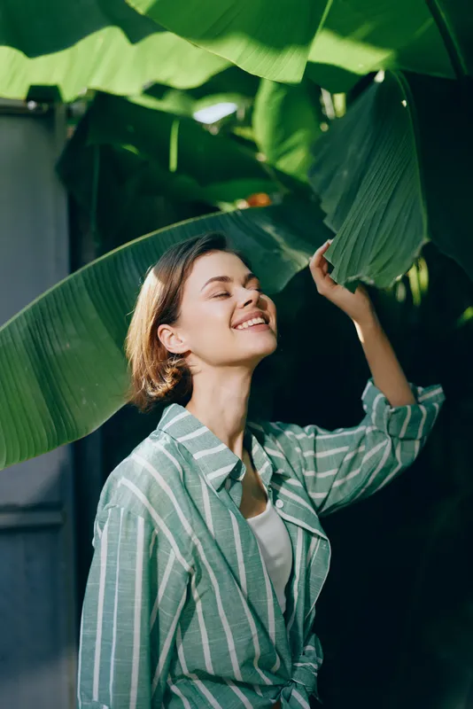 A joyful portrait of a woman among large tropical leaves, wearing a striped shirt, with a bright, carefree smile.